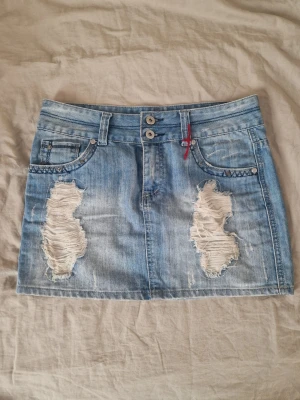 Ljusblå jeanskjol med slitningar -  Ljusblå jeanskjol med dubbla knappar och råa slitningar framtill med snygga detaljer på bakfickorna. Perfekt för en avslappnad och cool stil. Knapparna är lite lösa men funkar fortfarande utmärkt. Midjemåttet tvärs över är 40 cm - längden är 33.5 cm 