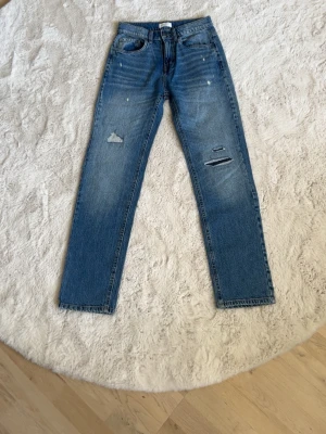 Blå raka jeans med slitningar - Säljer ett par blå jeans med raka ben med slitningar på både fram- och baksida. Jeansen har klassisk femficksmodell och är tillverkade i denim. 