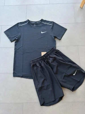 Svart träningsset från Nike - Snyggt svart träningsset från Nike med en kortärmad t-shirt och matchande shorts. Båda plaggen har Nike-loggan i vitt och är tillverkade i ett lätt och snabbtorkande material som passar perfekt för löpning eller gym. T-shirten har diskreta ränder och shorts med elastisk midja.
