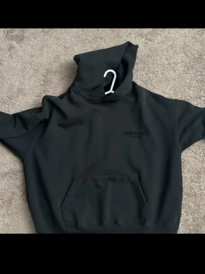 Svart Essentials Fear of God hoodie - Svart hoodie från Essentials Fear of God med stor logga på ryggen och mindre tryck på bröstet. Klassisk känguruficka framtill och stor huva. Tillverkad i mjukt bomullsmaterial, perfekt för en avslappnad streetwear-look.