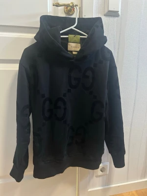 Svart hoodie från Gucci med GG-mönster - Säljer en svart hoodie från Gucci med stora GG-loggor i sammet på hela tröjan. Hoodien har en klassisk huva och är tillverkad i mjukt material. Snygg och lyxig streetwear-vibe som passar perfekt till jeans eller joggers.