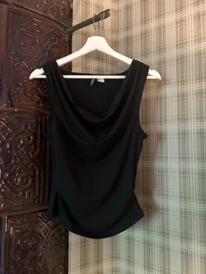 Svart draperad ärmlös topp H&M - Snygg svart ärmlös topp från H&M med draperad urringning och mjukt, stretchigt tyg. Perfekt för dig som gillar en stilren och trendig look. Passar till både jeans och kjol för en cool outfit.