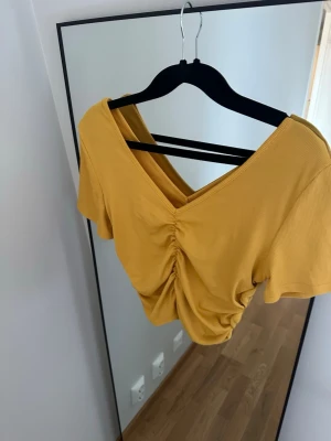 Gul croppad topp med snörning - Trendig gul croppad topp med v-ringning och snygg snörningsdetalj framtill. Toppen har korta ärmar och är tillverkad i ett ribbat material som ger en skön och stretchig passform. Perfekt för dig som vill ha en färgklick i garderoben.