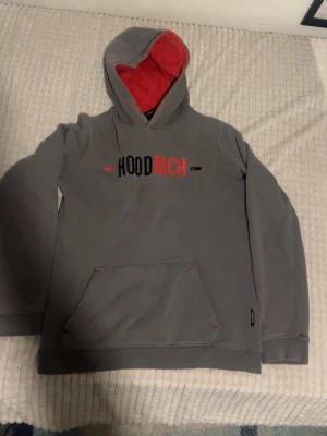Grå HOODRICH hoodie med röd detalj - Snygg grå hoodie från HOODRICH med röd insida i huvan och tryck i svart och rött på bröstet. Stor känguruficka framtill och logga även på ryggen. Perfekt för en avslappnad streetwear-look. Tillverkad i mjukt material för extra komfort.