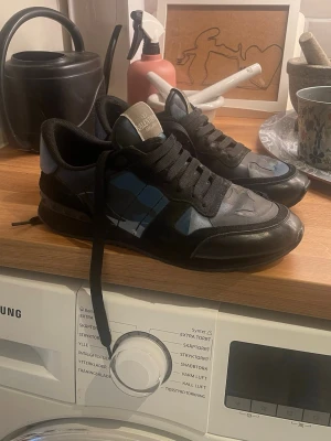 Svarta och blå sneakers från Valentino - Snygga sneakers från Valentino i svart och blått med coola detaljer och svarta skosnören. Skorna har en låg siluett och är tillverkade i en mix av läder och textil. Perfekta för dig som vill ha en stilren men ändå unik look. Säljer för dom va för små