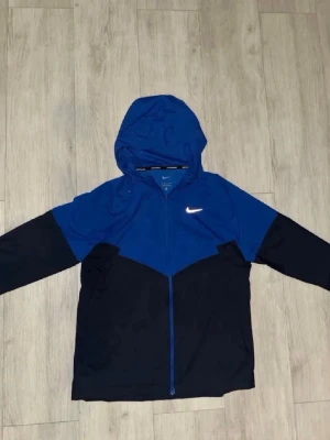 Blå och svart Nike vindjacka - Snygg vindjacka från Nike i blått och svart med hel dragkedja framtill och huva. Jackan har en sportig look med den klassiska Nike-loggan på bröstet. Perfekt för blåsiga dagar och enkel att matcha med streetwear. Perfekt inför sommaren 