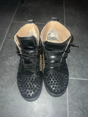 Svarta Christian Louboutins med nitar - Unika svarta sneakers med högt skaft och svarta nitar över hela skon. Skorna har snörning och detaljer i läderimitation samt beige insida. Perfekta för dig som vill sticka ut med en edgy stil.