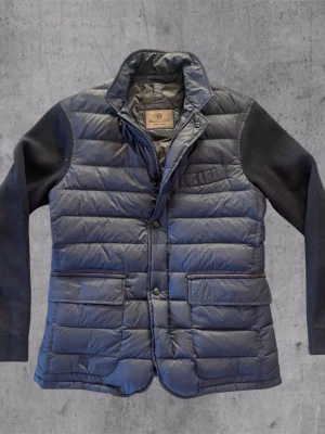 Mörkblå dunväst från Massimo Dutti - Snygg mörkblå dunväst från Massimo Dutti med quiltad design och stickade svarta ärmar. Västen har hög krage, dragkedja och tryckknappar framtill samt flera fickor. Perfekt för lager på lager och en stilren look.