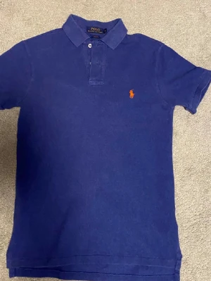 Blå pikétröja från Ralph Lauren - Klassisk blå pikétröja från Ralph Lauren med orange broderad logga på bröstet. Slim fit-modell med korta ärmar och krage med knappar. Perfekt för en clean och stilren look.