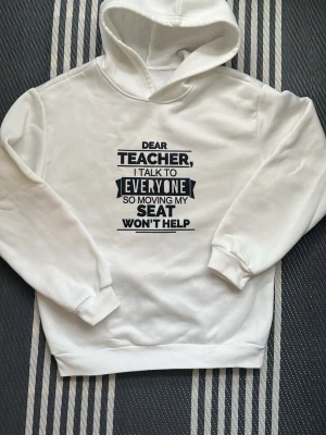 Vit hoodie med texttryck - Snygg vit hoodie med svart texttryck på bröstet: 'Dear teacher, I talk to everyone so moving my seat won't help'. Hoodien har huva och ribbade muddar vid ärmar och nederkant. Perfekt för dig som gillar statement-plagg och vill sticka ut i mängden. Storlek 160.