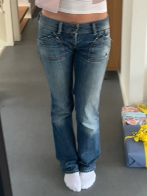 Blå bootcut jeans med låg midja - Säljer ett par blå bootcut jeans med låg midja och slitningar framtill. Jeansen har framfickor med snedskärning och klassiska bakfickor. Perfekta för en avslappnad och trendig look. Lite vinted och slitig stil, riktigt snygga! 