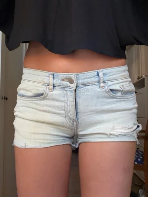 Ljusblå denimshorts med slitningar - Snygga ljusblå jeansshorts i klassisk femficksmodell med slitna detaljer och råa kanter. Shortsen har hög midja och dragkedja med knapp framtill. Perfekta för varma dagar och en avslappnad look.