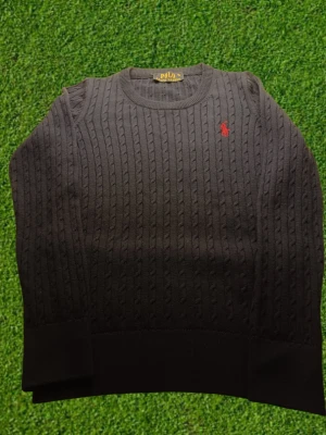 Mörkblå stickad tröja från Polo Ralph Lauren - Mörkblå stickad tröja från Polo Ralph Lauren med klassiskt kabelstickat mönster och röd broderad logotyp på bröstet. Rund halsringning och långa ärmar. Perfekt för dig som gillar stilrena och tidlösa plagg.