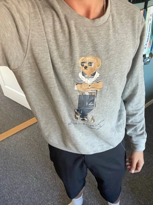 Grå sweatshirt Polo Bear Ralph Lauren - Säljer en grå sweatshirt från Ralph Lauren med den ikoniska Polo Bear-figuren tryckt på bröstet. Tröjan har rund halsringning och är tillverkad i mjukt bomullsmaterial. Perfekt för en avslappnad och stilren look.