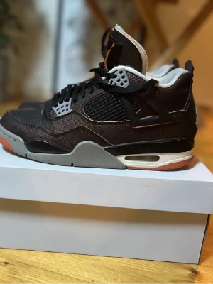 Svarta Air Jordan 4 sneakers - Säljer ett par svarta Air Jordan 4 sneakers med meshpaneler, grå och orange detaljer samt vita inslag. Skorna har en ikonisk siluett med synlig Air-enhet i sulan och klassiska plastdetaljer vid snörningen. Perfekta för dig som gillar streetwear och snygg design. Kvinnomodell men passar unisex