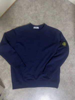 Mörkblå sweatshirt från Stone Island - Mörkblå sweatshirt från Stone Island med klassisk rund halsringning och ribbade muddar. Tröjan har den ikoniska Stone Island-loggan på vänster ärm. Perfekt för en avslappnad och stilren look.