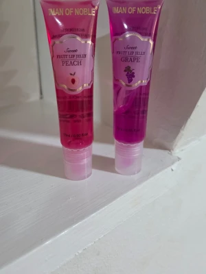 Fruit Lip Jelly  - Säljer två läppglans från IMAN OF NOBLE i smakerna Peach och Grape. Ena tuben är persikorosa och den andra är lila, båda i genomskinlig plast med skruvkork. Ger glansig finish och har fruktig doft. Perfekt för dig som vill ha mjuka och glansiga läppar med en hint av färg.