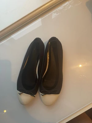 Svarta ballerinaskor med beige tå - Stilrena ballerinaskor i svart tyg med beige tå och liten rosett framtill. Klassisk rundad form och låg sula, perfekta för en enkel och snygg look. Passar till många olika outfits och är lätta att matcha. Endast använda en gång! (Små skador/repor som syns på sista bilden)