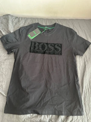 Hugo boss t-shirt - Snygg svart t-shirt från Hugo Boss med stor logga framtill dekorerad med svarta strassdetaljer. Klassisk rund halsringning och korta ärmar. Perfekt för dig som vill ha en stilren men ändå iögonfallande look.