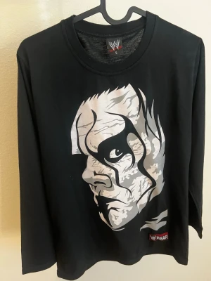 Svart WWE långärmad topp Sting - Svart långärmad topp från WWE med stort tryck av Sting på framsidan och STING-tryck i vitt på ryggen. Tillverkad i mjuk bomull och har en normal passform. Perfekt för wrestlingfans som vill ha en unik och snygg topp.