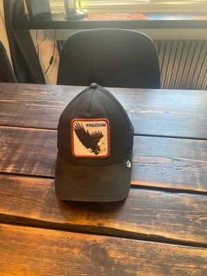 Svart truckerkeps med örnpatch - Säljer en svart truckerkeps från Goorin Bros med mesh på sidorna och bak. Framtill sitter en patch med en örn och texten 'FREEDOM'. Kepsen har böjd skärm och justerbar snapback. Perfekt för dig som gillar streetwear och coola detaljer.