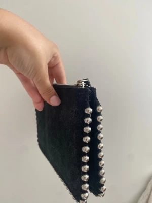 Svart kuvertväska med silverdetaljer - Stilren svart kuvertväska i mockaliknande material med coola silvriga kulor längs kanterna. Perfekt att bära i handen och stängs med dragkedja upptill. Enkel men edgy design som lyfter vilken outfit som helst.