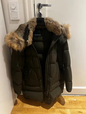 Mörkgrön pufferjacka från Cedrico - Snygg mörkgrön pufferjacka från Cedrico med stor fluffig päls på huvan och perfekt för de kalla dagarna nu för vintern. Jackan har dragkedja framtill, flera fickor med dragkedja och snörning nedtill och har en riktigt schysst oversized look. Skriv efter minsta lilla fundering!!