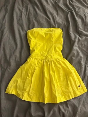 Gul axelbandslös klänning Hollister - Superfin gul axelbandslös klänning från Hollister med kort kjol och markerad midja. Klänningen har en enkel design med liten broderad logga nertill och är perfekt för varma dagar. Sitter som xs
