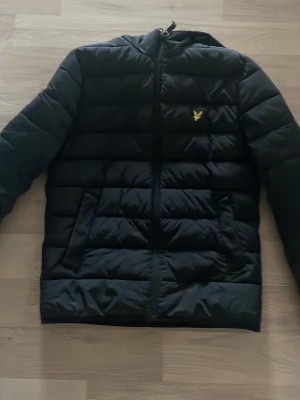 Svart pufferjacka från Lyle & Scott - Säljer en svart pufferjacka från Lyle & Scott med gul logga på bröstet. Jackan har dragkedja framtill, två sidofickor och en klassisk quiltad design. Perfekt för kalla dagar och riktigt skön att bära. Materialet är syntet och jackan har en modern passform. Priset kan diskuteras vid snabbaffär 