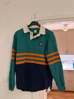 Adidas rugbytröja med ränder - Retroinspirerad långärmad rugbytröja från Adidas med grön överdel, marinblå nederdel och orange ränder över bröst och ärmar. Vit krage och knappslå framtill. Adidas-logga broderad på bröstet. Perfekt för en sportig och avslappnad stil.