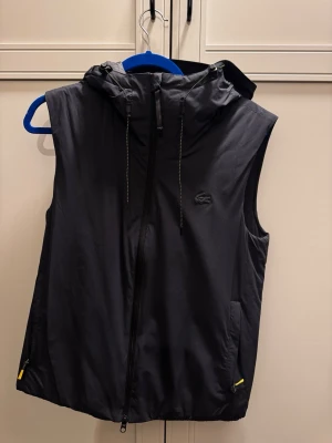 Svart väst med huva från Lacoste - Snygg svart väst från Lacoste med dragkedja framtill, huva och två fickor med gula detaljer på dragkedjorna. Västen har en minimalistisk look med Lacoste-loggan på bröstet och är tillverkad i ett lätt, sportigt material.