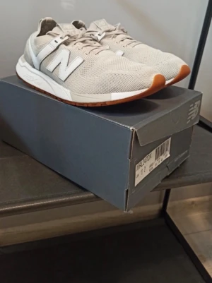 Beige New Balance 247 sneakers - Snygga beige New Balance 247 sneakers med vit sula och gummidetaljer på tån. Ovandelen är i mesh för bra andningsförmåga och komfort. Klassisk N-logga på sidan och diskreta detaljer i vitt. Perfekta för en stilren och sportig look.