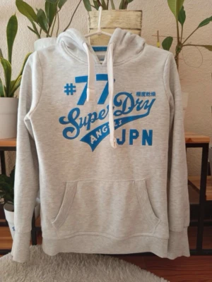Grå Superdry hoodie med blått tryck - Snygg ljusgrå hoodie från Superdry med stort blått tryck på bröstet och vita dragsnören i huvan. Klassisk känguruficka framtill och mjukt material som känns skönt mot huden. Perfekt för en avslappnad streetwear-look.