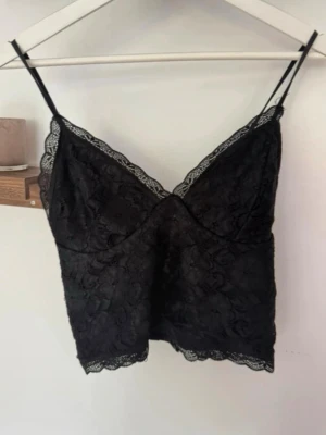 Svart spetslinnetopp med tunna band - Säljer en svart linnetopp med tunna axelband och vacker spetsdetalj längs kanterna. Toppen har en feminin v-ringning och är tillverkad i ett mjukt spetsmaterial som ger en elegant look. Perfekt att styla med jeans eller kjol.