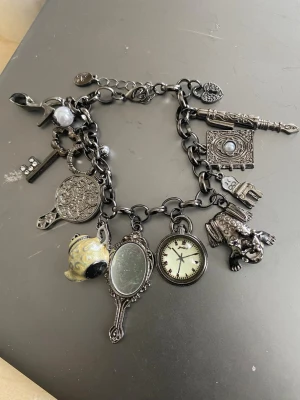 Chunky charmarmband med printed klocka - Unikt chunky armband i mörk metall med flera detaljerade berlocker som spegel, klocka, tekanna, nyckel, pärla och djur. Armbandet har vintagekänsla och cool mix av former och material, inklusive emalj och glas. Perfekt för dig som gillar statement-smycken och personlig stil. its a printed clock
