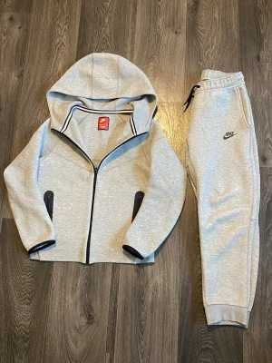 Grå Nike hoodie med dragkedja - Snygg ljusgrå hoodie från Nike med hel dragkedja framtill och svarta detaljer vid fickorna och muddarna. Stor huva och två praktiska fickor med dragkedja. Perfekt för en sportig och avslappnad stil. Tillverkad i mjukt och bekvämt material.