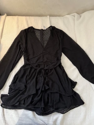 Svart kort klänning från Amisu - Snygg svart kort klänning från Amisu med lång ärm, v-ringning och omlottkänsla. Klänningen har volangdetaljer och knyt i midjan. Tunt, lätt material med diskret prickigt mönster och dragkedja bak. Perfekt för dig som gillar trendiga och feminina plagg.