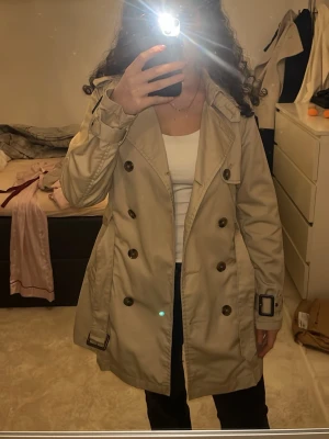 Beige trenchcoat med bälte och knappar - Stilren beige trenchcoat med dubbelknäppning, bälte i midjan och dekorativa spännen vid ärmslut. Klassisk krage och axelklaffar. Perfekt för lager-på-lager och snyggt till jeans eller klänning. Har en liten fläck