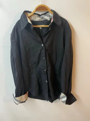 Svart skjorta från Burberry - Stilren svart skjorta från Burberry i storlek M men passar Xs. Klassisk krage och knäppning framtill, med ikoniskt rutigt mönster på insidan av krage och manschetter. Tillverkad i mjuk bomull som känns skön mot huden. Perfekt för dig som gillar en clean och tidlös look.