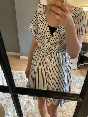 Vit och svart randig omlottklänning - Snygg kort omlottklänning i vitt med svarta ränder. Klänningen har volangdetaljer, knyt i midjan och v-ringning. Korta ärmar och lätt, luftig känsla, perfekt för varma dagar. Använd en gång, ej bra pris,,, skriv till mig