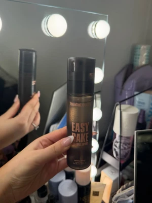 Huda Beauty Easy Bake Setting Spray - Face mist från Huda Beauty i svart och bronsfärgad flaska. Easy Bake Setting Spray ger en fräsch och återfuktande känsla samt fixerar sminket. Praktisk sprayflaska i plast, perfekt att ha i väskan för touch-ups under dagen.