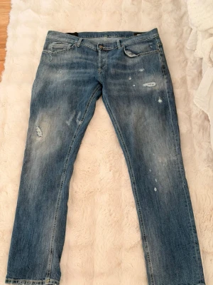 Blå slitna jeans med orange detalj - Säljer ett par blå jeans med slitningar och distressed detaljer. Jeansen har raka ben och klassisk femficksmodell. På baksidan finns en orange broderad detalj vid linningen och en unik söm på bakfickan. Perfekta för en avslappnad streetstil.