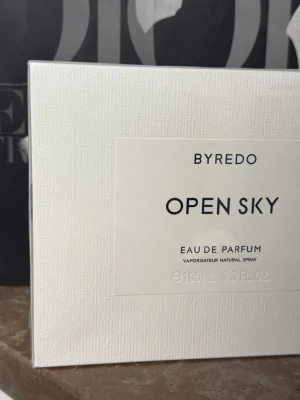 Byredo Open Sky Eau de Parfum 100ml - Byredo Open Sky Eau de Parfum i elegant vit kartong med subtilt mönster. Förpackningen har en stilren design med svart text och innehåller 100 ml doft. Perfekt för dig som gillar exklusiva och moderna parfymer.