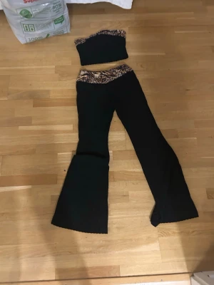 Svart set med leoparddetaljer - Tvådelat set med svarta utsvängda byxor och matchande tubtop. Båda delarna har leopardmönstrade detaljer i midjan och överdelen. Perfekt för dig som gillar trendiga och djurmönstrade plagg.