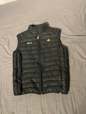 Svart dunväst från Ellesse - Snygg svart dunväst från Ellesse med quiltad design och hög krage. Västen har dragkedja framtill och logga på bröstet. Perfekt att slänga över en hoodie för extra värme. Klassisk och stilren look som funkar till det mesta.knappt använd