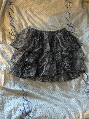 Svart volangkjol med prickigt mönster - Säljer en svart kortkjol med vita prickar och flera lager volanger. Kjolen har en elastisk midja och ett lekfullt, flowy utseende som passar perfekt till en trendig vardagslook.