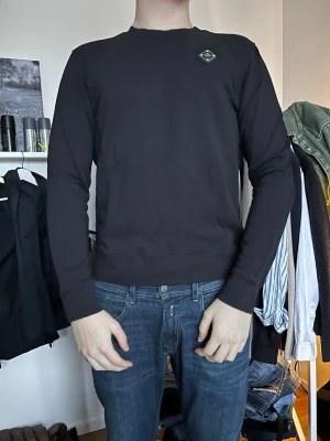 Svart sweatshirt från J.LINDEBERG  - Säljer en svart sweatshirt från Les Deux med rund halsringning och diskret logomärke på bröstet. Tröjan har långa ärmar och ribbade muddar vid ärmslut och nederkant. Perfekt för en clean och stilren look.