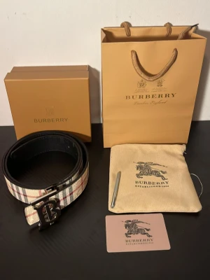 Burberry svart och rutig skinnbälte - Säljer ett exklusivt Burberry bälte i svart skinn med klassiskt rutigt mönster på insidan och en stor metallspänne med Burberrys logotyp. Kommer med originalpåse, box och shoppingpåse. Perfekt accessoar för att lyfta din outfit.
