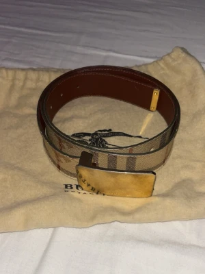 Burberry bälte - Beiget burberry bälte med guldigt spänne gjort i läder. köpte det på vestiaire collective så det är äkta kan nog ordna kvitto. Det är 2,5cm tjockt och 100 cm långt. Dustbag med kommer väldigt fint skick på bältet. Bara höra av sig om man undrar något eller skulle vilja ha fler bilder 