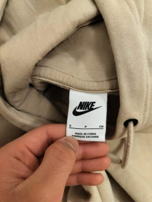 Beige hoodie från Nike med huva - Säljer en stilren beige hoodie från Nike med huva och dragsko. Hoodien har en klassisk passform och är tillverkad i mjukt material som känns skönt mot huden. Perfekt för en avslappnad look och enkel att matcha med andra plagg.